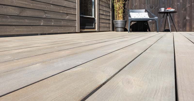 Nærbilde av grå terrassebord med hytta og hagemøbler i bakgrunn.