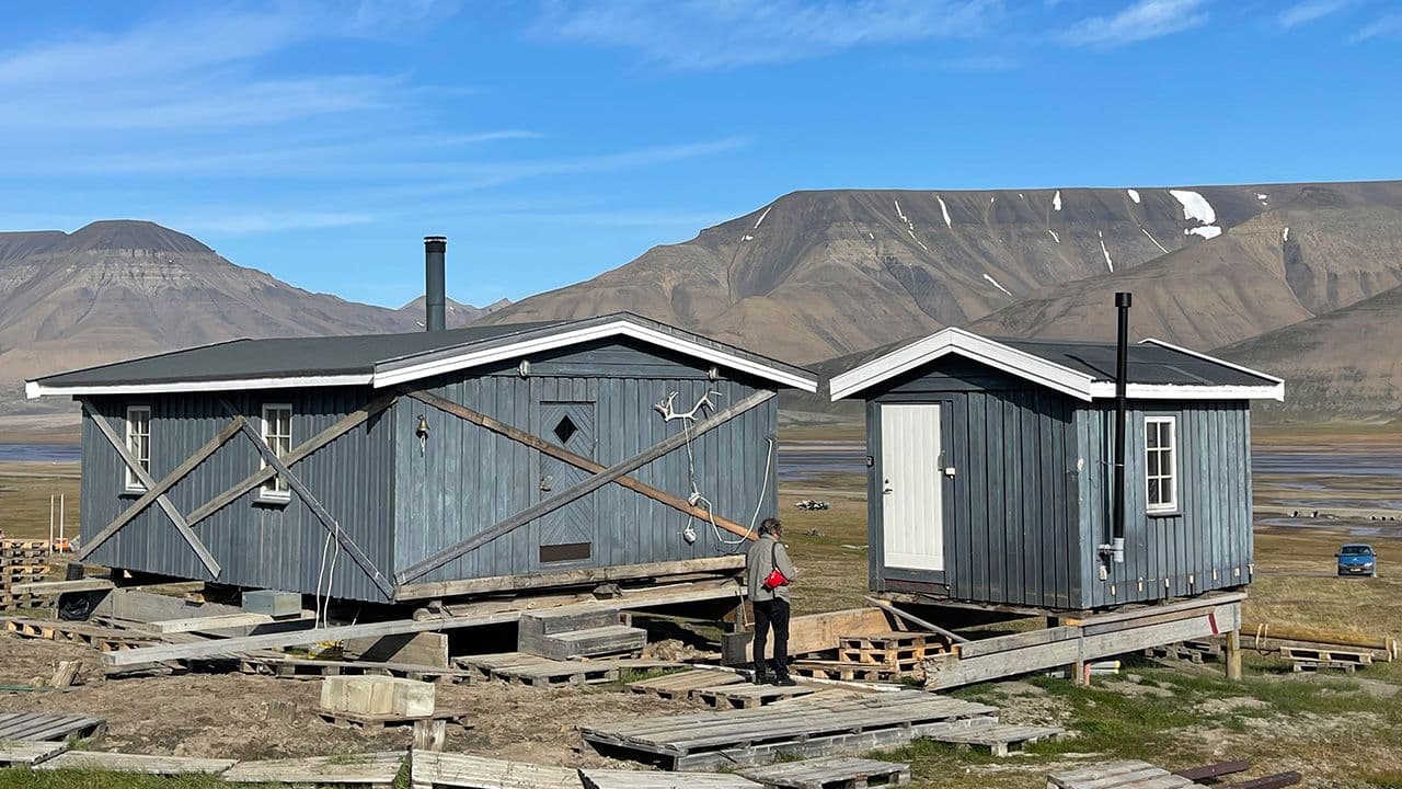 Blå hytte med tilbygg ute i naturen med en mann i forkant og fjell bak.