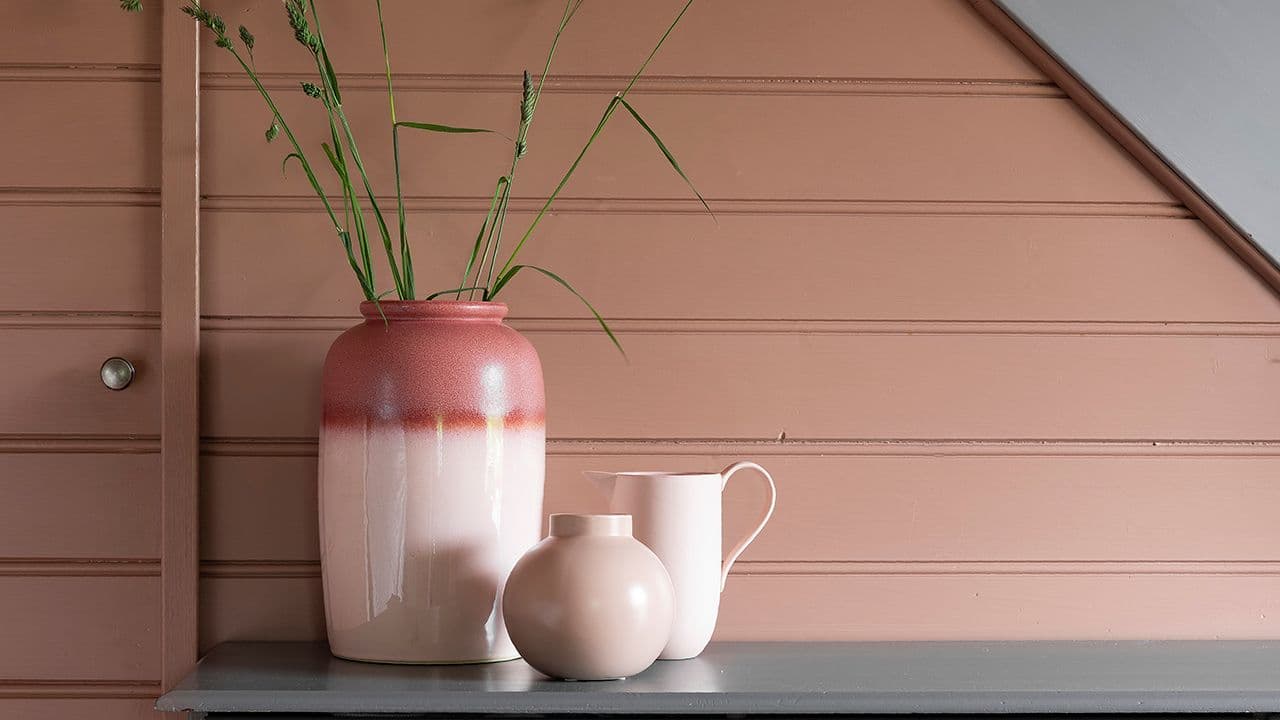 Nærbilde av liggende trepanel som er malt i en dus rosafarge. Foran står en gråmalt kommode med vaser og blomster på.